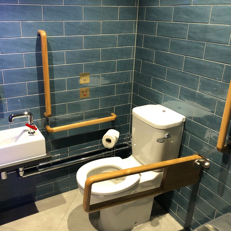 Ground Floor Accessible Toilet - Wooden Grab Rails at white toilet & sink. Large space for wheelchair. Right hand bar retracts & toilet 48cm high. Emergency Buttons on wall & floor. Sink at wheelchair height within reach of toilet Pale grey tiled flooring. Dark blue tiled walls. Well lit. Near Bar/Restaurant & Lifts