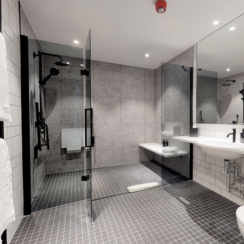 Liberty Wheelchair Access Room - Bathroom - Flooring is black non slip tiles. Pale grey walls. Removable support rails & shower seat. 3 grab rails in shower. 2 retractable & horizontal to shower seat Ceiling & hand held shower. Central shower post reinforced as grab rail. Fire emergency light on ceiling & call button in shower.