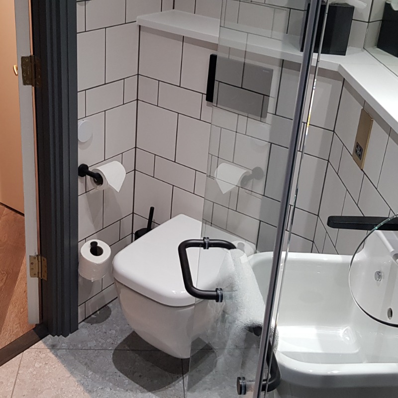 Liberty Ambulant Access Room - WC - 3"/5cm step into bathroom. Smooth non slip floor. Toilet 48cm high. No grab rails at WC. Grey Tiled floor & white tiled wall. Sink near WC & at standing height. Glass shower door porthole accesses temp. controls prior to shower. Black grab rail on shower door. Black taps on white good colour contrast.