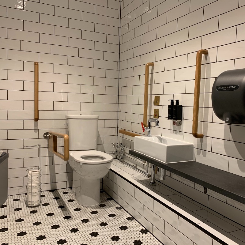 9th Floor Salvation Bar Accessible Toilet - Located behind Bar. Space to move wheelchair. 5 Brown wood grab rails around toilet & sink. Retractable rail for left hand transfer to toilet. Low sink within reach of toilet. 2 Emergency buttons on wall next to toilet & under sink. Patterned black & white non slip floor. White tiled walls & well lit.